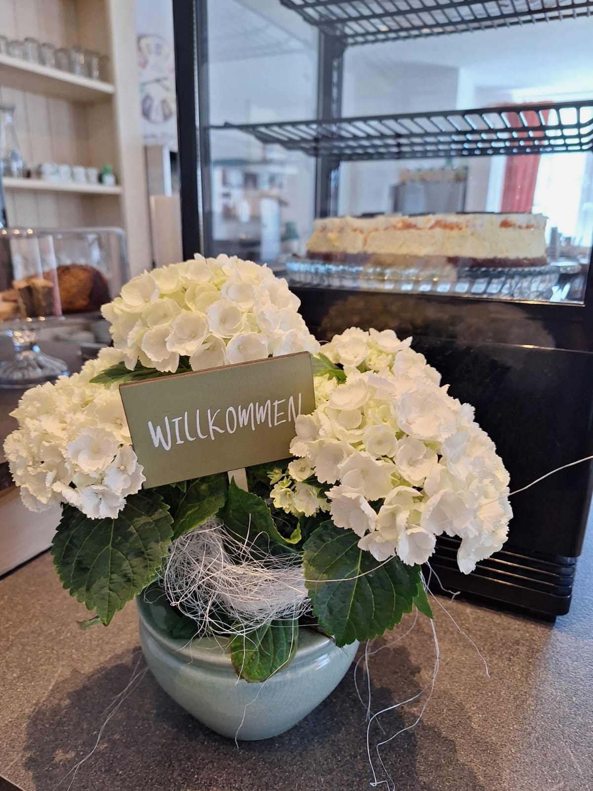 Willkommen-Schild mit weißen Hortensien an der Kuchentheke